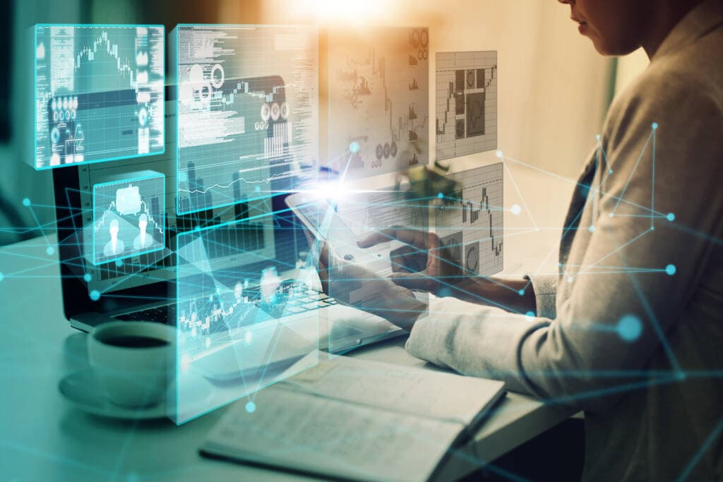 A person sits at a desk, immersed in application modernization on their laptop, surrounded by floating holographic screens showcasing graphs, charts, and data analytics. The desk features an open notebook, a cup of coffee, and a smartphone, creating an undeniably high-tech work environment.